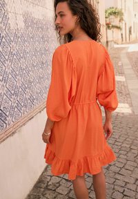 Orange, knälång klänning med puffärmar och volangkant; tillverkad av lätt tyg. Har en åtsittande midja och en enkel design.