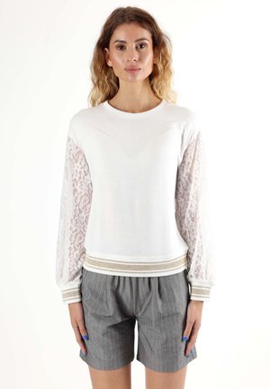 Le mannequin se tient face à l’avant, vêtu d’un haut blanc à manches en dentelle et à poignets rayés, associé à un short gris à fines rayures.