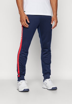 Hombre que lleva pantalones de chándal azul marino con una franja lateral roja y texto blanco "Lacoste", combinados con zapatillas blancas y una camisa blanca.