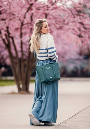 Junge Frau in blauem Rock, weiß-blau gestreiftem Pullover, weißen Sneakers und grüner Handtasche geht nahe blühenden rosa Kirschbäumen.