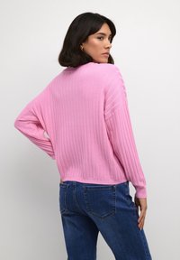 Roza rebrasta jopica z dolgimi rokavi, okroglim izrezom in ohlapnim krojem, v kombinaciji z modrimi denim hlačami. Gladka tekstura, ležeren dizajn.