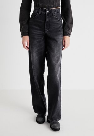 Persoon draagt zwarte jeans met hoge taille en wijde pijpen met vervaagde details, gecombineerd met zwarte laarzen en een korte zwarte spijkerjas.