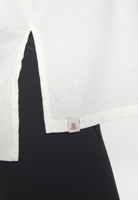White short-sleeved top with side slits and a branded tag at the hem. The fabric appears soft, and it's paired with black leggings.