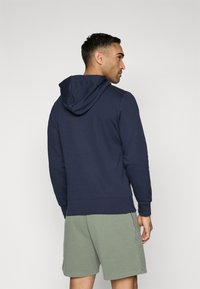 Sweatshirt azul-marinho com capuz e corte relaxado, apresentando mangas longas e um bolso tipo canguru, combinado com calções verde-claro.