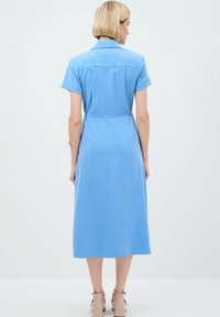 Robe midi bleu clair avec des manches courtes, un col de chemise, un détail de empiècement dans le dos et une texture lisse. Assortie à des chaussures à talons beige.