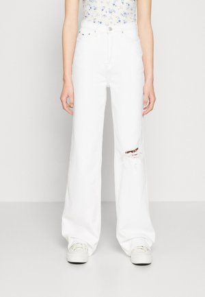 Femme portant un jean blanc taille haute à jambes larges avec une déchirure sur le genou droit et des baskets blanches, debout devant un fond clair uni.