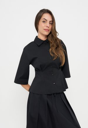 Femme aux longs cheveux bruns portant une chemise noire à boutons avec des manches jusqu'aux coudes et une jupe noire assortie sur un fond uni.