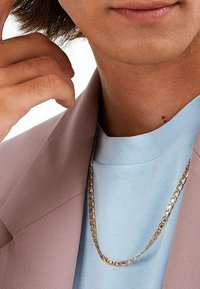 Goldene Kettenhalskette mit flachen, miteinander verbundenen rechteckigen Gliedern. Über einem hellblauen Hemd getragen, teilweise von einem pastellrosa Blazer verdeckt.