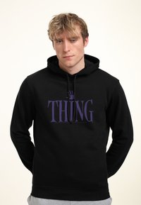 Felpa nera con grafica "THING" viola, cappuccio con cordino e tasca frontale. Il modello è in piedi contro uno sfondo chiaro, indossando pantaloni grigi.