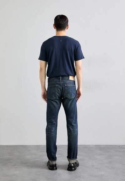 Dunkelblaues T-Shirt kombiniert mit dunklen Jeans. Das Shirt hat kurze Ärmel und ein kleines Detail auf der Rückseite, während die Jeans mit gesteppten Taschen ausgestattet sind.