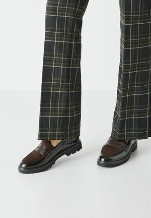 Braune Loafers mit einem glänzenden schwarzen Absatz, ausgestattet mit einem strukturierten braunen Paneel und zwei horizontalen Riemen. Getragen mit karierter, weit geschnittener Hose.