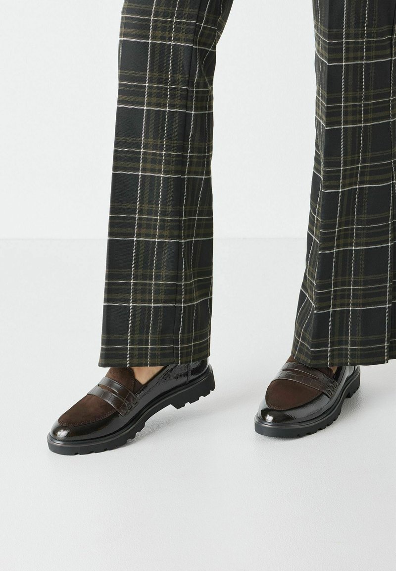 Brown loafers with a shiny black heel, featuring a textured brown panel and two horizontal straps. Worn with plaid wide-leg trousers.