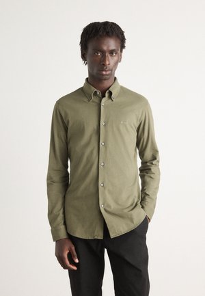 Camisa de manga larga de color verde oliva con cuello abotonado, botones frontales y bordado de logo discreto, combinada con pantalones oscuros.