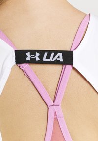 Bílá sportovní podprsenka s růžovými ramínky; má černou štítek s logem "UA". Ramínka mají překřížený design a hladkou texturu.
