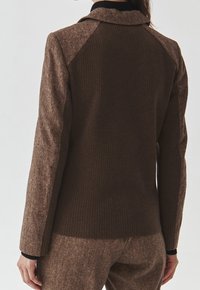 Ruskea tweed-takki, jossa on ribbineulottu pinta, kontrastivärinen kangas olkapäillä, istuva malli ja lovettu kaulus. Sileä, jäsennelty siluetti.