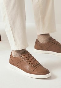 Bruna lädersneakers med perforerade sidopaneler, rund tå och vita gummisulor, i kombination med beige byxor och texturerade strumpor.