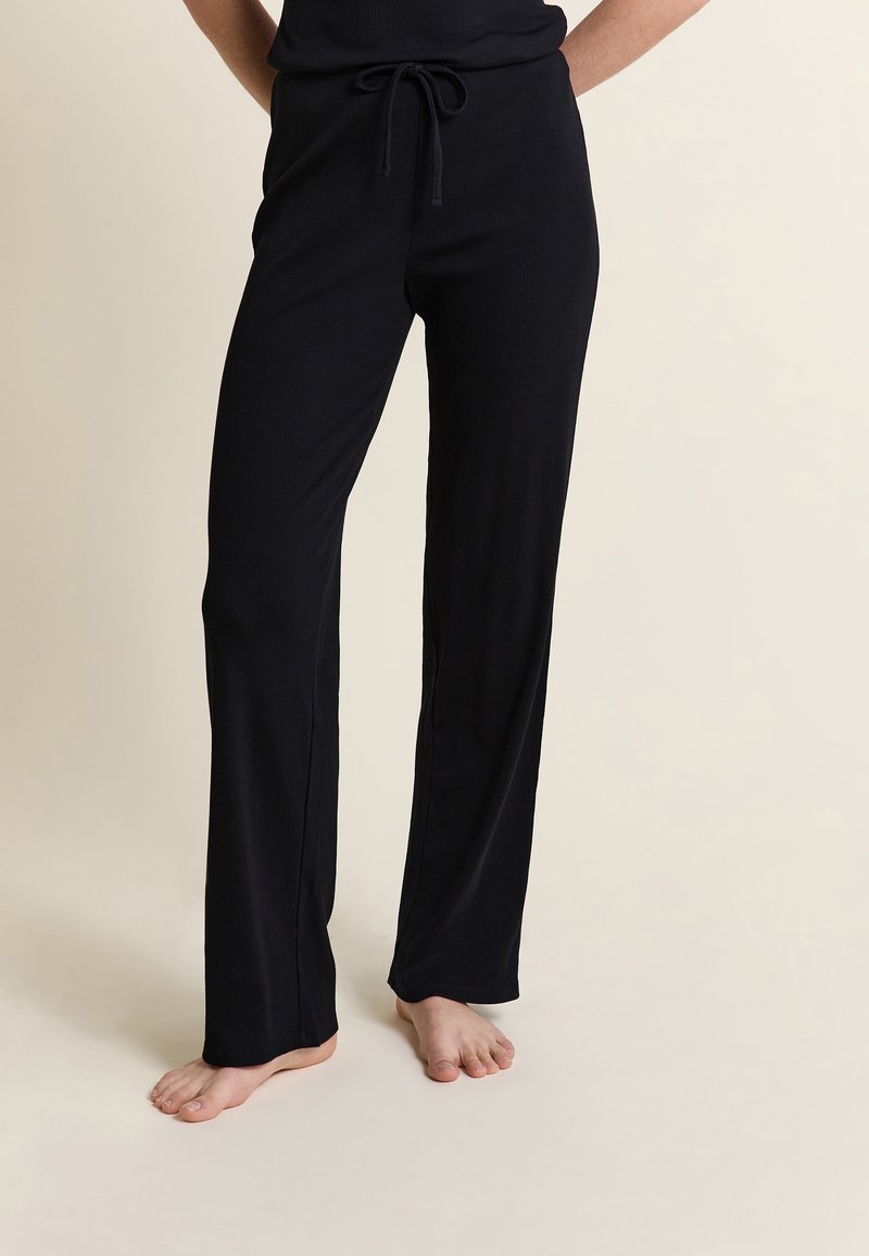 Persona vistiendo pantalones de chándal negros con cordón, de pie descalza sobre un suelo y fondo de color claro.