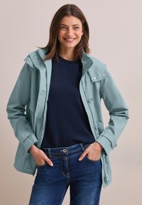 Hellpetrolfarbene Jacke mit Kapuze, Druckknöpfen und Seitentaschen, getragen über einem marineblauen Hemd, kombiniert mit dunklen Jeans aus Denim. Glatte Stofftextur.