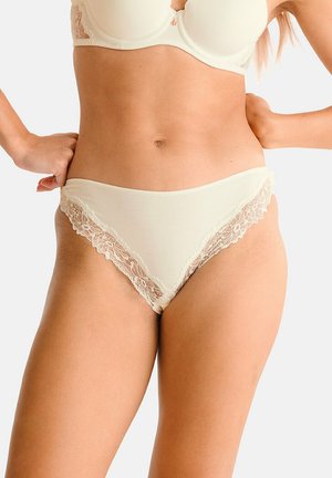 Vrouw die crèmekleurig ondergoed met kanten rand en bijpassende beha draagt, handen op de heupen, tegen een effen lichte achtergrond.