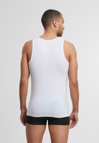Top sin mangas blanco de algodón, con un diseño ajustado y cuello de canalé. El modelo lleva pantalones cortos negros, visto desde atrás.