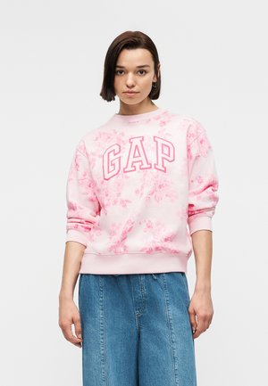 Młoda kobieta ubrana w różową kwiatową bluzę GAP i luźne niebieskie dżinsy, stojąca na tle jednolitego jasnego tła.