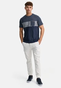 Camiseta bloqueada en color marino y gris con un bolsillo, combinada con pantalones cargo claros. El modelo lleva zapatillas oscuras, creando un aspecto casual.