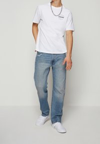 Bílý krátký rukáv tričko s černým kruhovým logem, spárované s světle modrými rovnými džíny a bílými teniskami; jednoduchý, casual design.