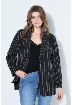 Kobieta z luźnymi falującymi włosami, ubrana w czarną marynarkę w prążki, czarny top zdobiony koronką, niebieskie dżinsy i pierścionki na palcach.