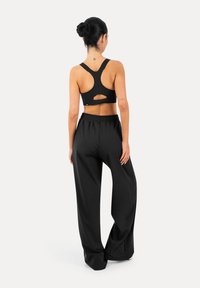 Frau mit dunklem Haar im Dutt, die einen schwarzen Racerback-Sport-BH und lockere schwarze Hosen trägt, steht mit dem Rücken zum Betrachter.