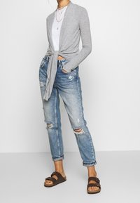 Ljusgrå stickad cardigan knuten i midjan, tillsammans med slitna, ljusblå denimjeans och bruna sandaler. Enkla lager av halsband.