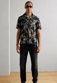 Camicia a maniche corte nera e verde con motivo floreale e colletto, abbinata a jeans di denim scuri e scarpe marroni.