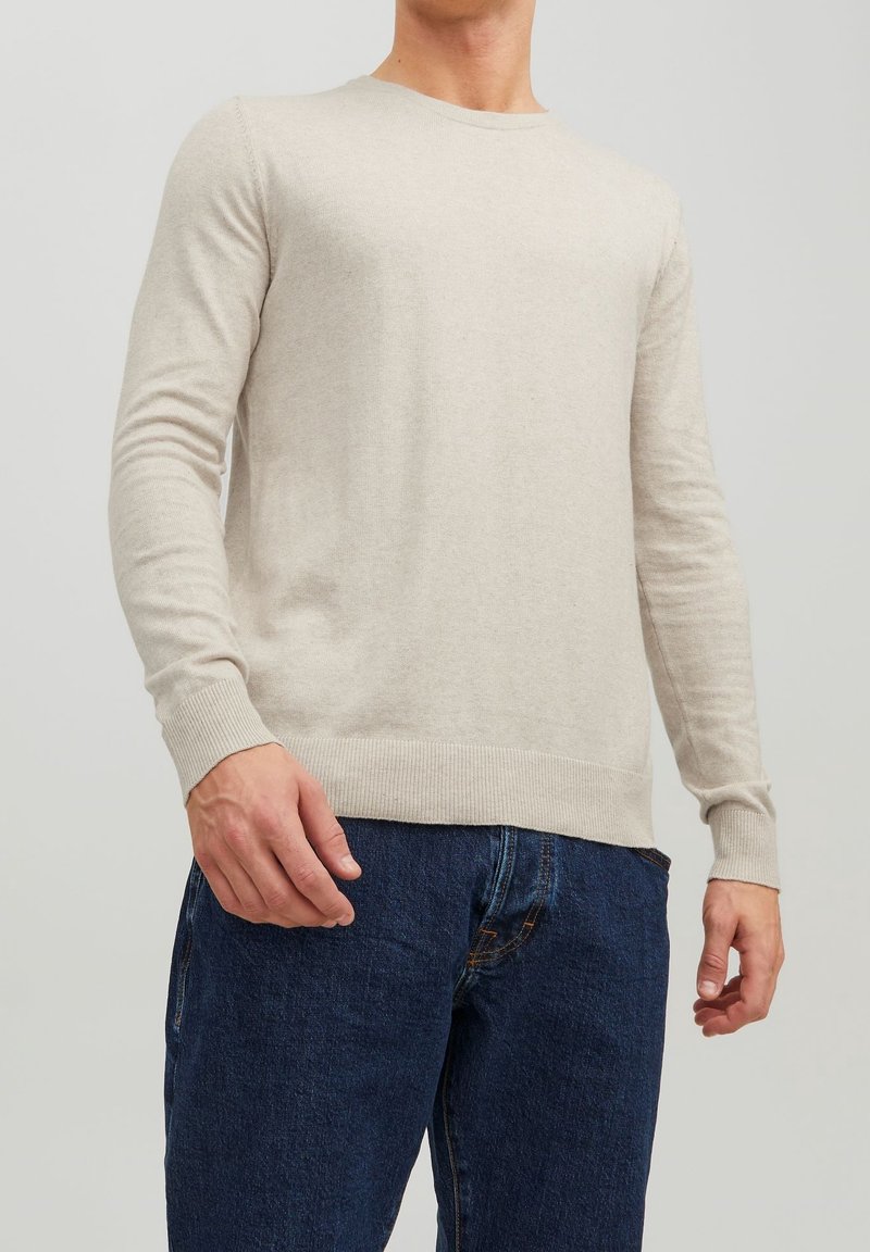Homme portant un pull beige à manches longues et un jean bleu foncé, debout devant un fond clair uni.