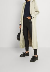 Beige long trench coat over navy long-sleeve top, black leather skinny pants, and black shoes with a bow detail. Black handbag present.