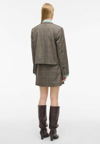 Bruine geruite blazer met een cropped ontwerp, gecombineerd met een bijpassende geruite rok. Model draagt hoge, slungelige bruine leren laarzen.
