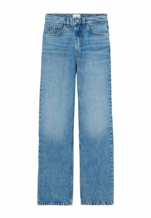 Donna in piedi che indossa una giacca di pelle marrone corta, una camicia scura a righe, jeans azzurri e stivaletti neri con punte appuntite.