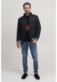 Svart puffjakke med horisontale sømmer, grå mansjetter og oransje glidelåsdragere. Brukt over en marineblå og grønnstripete polo og blå jeans.