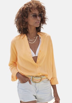 Light orange button-up shirt with rolled sleeves, worn over a white bikini top. Paired with blue and yellow woven belt and denim shorts.