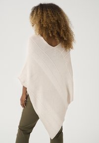 Beige gestrickter Poncho mit einem V-Ausschnitt und diagonaler Rippstruktur. Hat lockere Ärmel und eine lässige Passform, kombiniert mit olivgrünen Hosen.