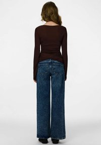 Brun långärmad topp med figursydd design och rynkdetalj, kombinerad med högmidjade vida blå jeans i denim och svarta skor.