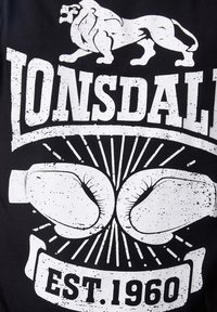 Zwart T-shirt met een witte verweerde graphic van een leeuw, bokshandschoenen en "LONSDALE" boven "EST. 1960," met stralende lijnen rondom de handschoenen.