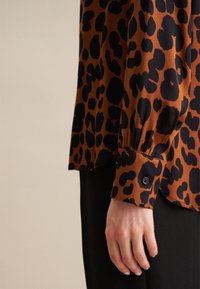 Blusa con stampa leopardata in arancione e nero, con maniche lunghe, polsini con bottoni e una consistenza liscia. Pantaloni scuri indossati sotto.
