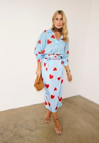 Γυναίκα που φοράει ανοιχτό μπλε μπλούζα και φούστα με κόκκινα σχέδια καρδιά, κρατώντας καφέ τσαντάκι, φοράει ψηλοτάκουνα παπούτσια με σχέδιο λεοπάρ και καφέ φουλάρι στο λαιμό.