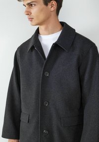 Dunkelgrauer Wollmixmantel mit Hemdkragen, Knopfverschluss und seitlichen Taschen. Glatte Textur mit leichtem Glanz.