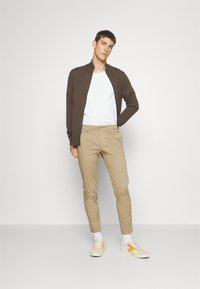 Bruine zip-up jas over een witte t-shirt, beige slim-fit broeken en lichte sneakers met gele accenten. Soepele textuur en casual stijl.