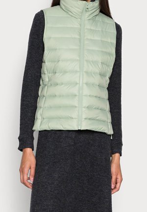 Kobieta ubrana w jasnozieloną pikowaną kamizelkę bez rękawów z zamkiem błyskawicznym na długim, ciemnoszarym sukience z długim rękawem, stoi z rękami wzdłuż ciała.