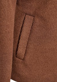Gros plan d'une poche de manteau en laine marron avec un rabat propre et rectangulaire et un détail de tissu texturé.