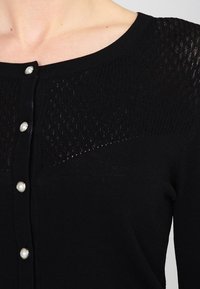 Černý cardigan vyrobený z měkké pleteniny s texturovaným vzorem na ramenech, se perlovými knoflíky po celé přední části.