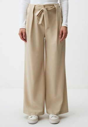 Pantaloni beige a gamba larga con vita legata, caratterizzati da una texture liscia e un design fluido. Indossati con sneakers bianche.