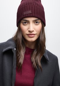 Gorro de punto burdeos con textura acanalada y un parche con logo, llevado con una chaqueta negra con cremallera sobre una camiseta burdeos.
