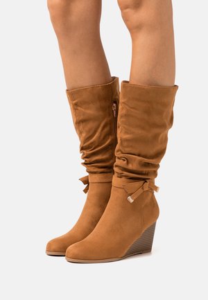 Botas de cuña de ante marrón con un diseño arrugado, cremallera lateral y detalle decorativo de lazo en el tobillo. Tacón de cuña para mayor altura.